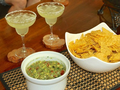 Chunky Guacamole in a deep bowl next to a bowl of tortilla chips and two margaritas
