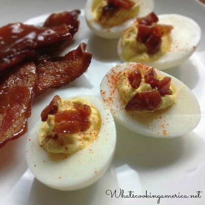 Deviled eggs with candied bacon on a white plate with two sliced of bacon on the side